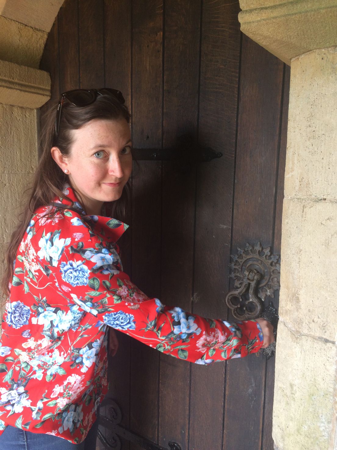 A women with brown long hair in a red patterned shirt is opening an old wooden door.