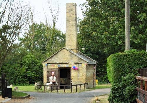 Person sat outside the open door of a small square stone structure with tall chimney surrounded by trees and bushes. 