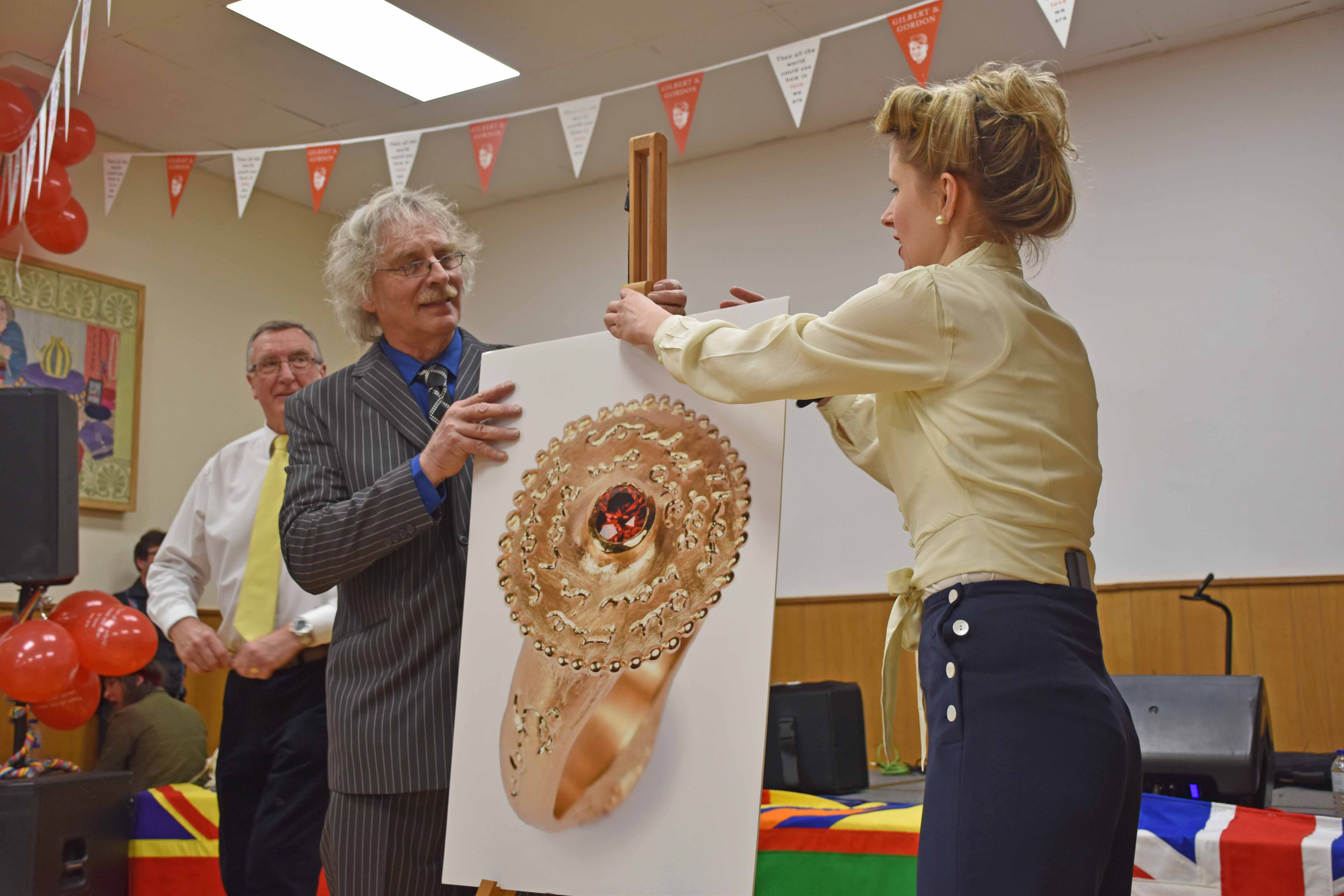 Older man and younger woman in 1940s outfit adjusting a poster of a gold ring with ruby centre.