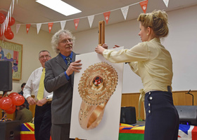 Older man and younger woman in 1940s outfit adjusting a poster of a gold ring with ruby centre.