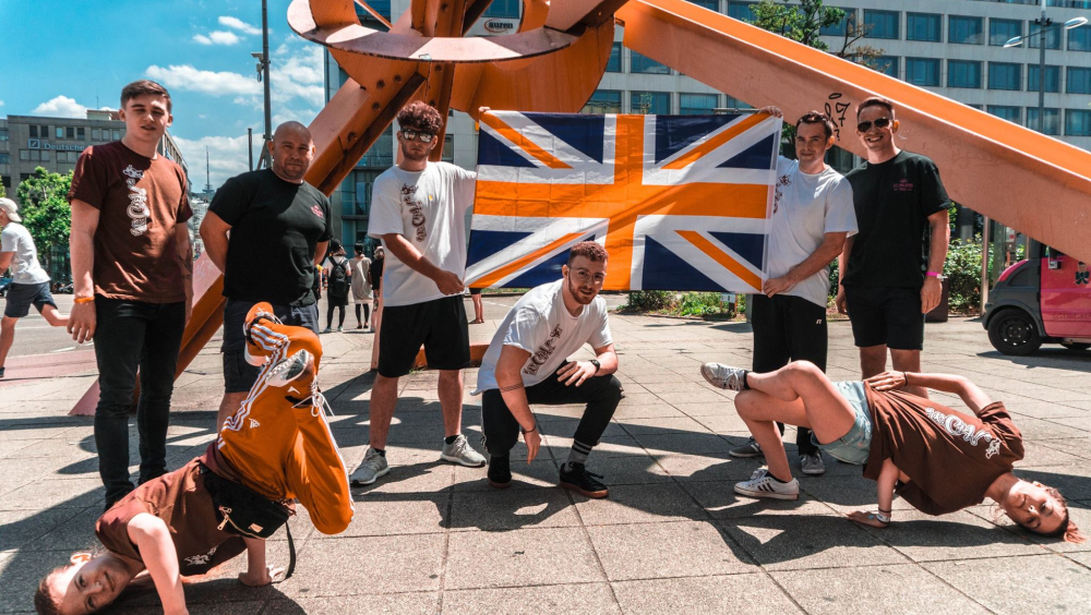 Group of young male breakdancers performing outside a modern building and orange metal sculpture. 