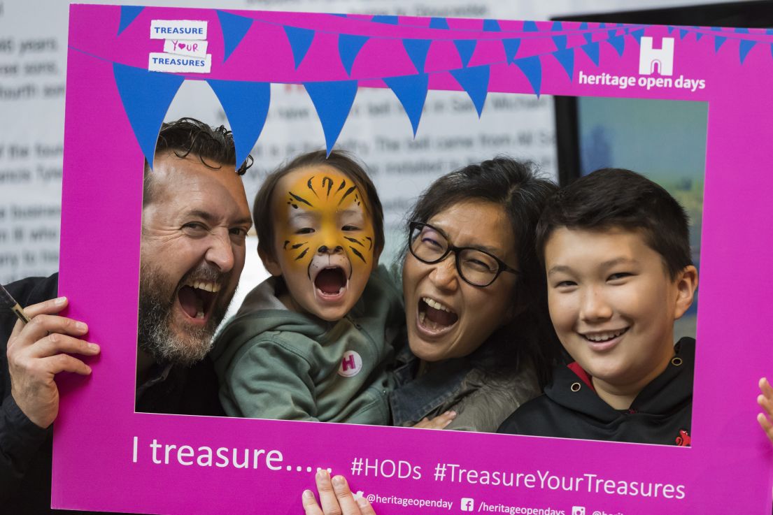 An image taken of a family with two small children, holding up a cardboard selfie frame and making fun faces towards the camera.