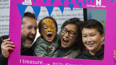 An image taken of a family with two small children, holding up a cardboard selfie frame and making fun faces towards the camera.