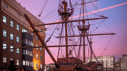 Masts of a sail boat in a city environment. The sky is pink & purple as dusk approaches.