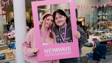 Two young adults looking through a pink selfie frame in a room decorated with bunting.