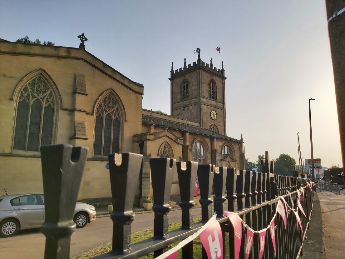 Pink bunting hung on metal railings in front of a stone church flying a flag from its tower.