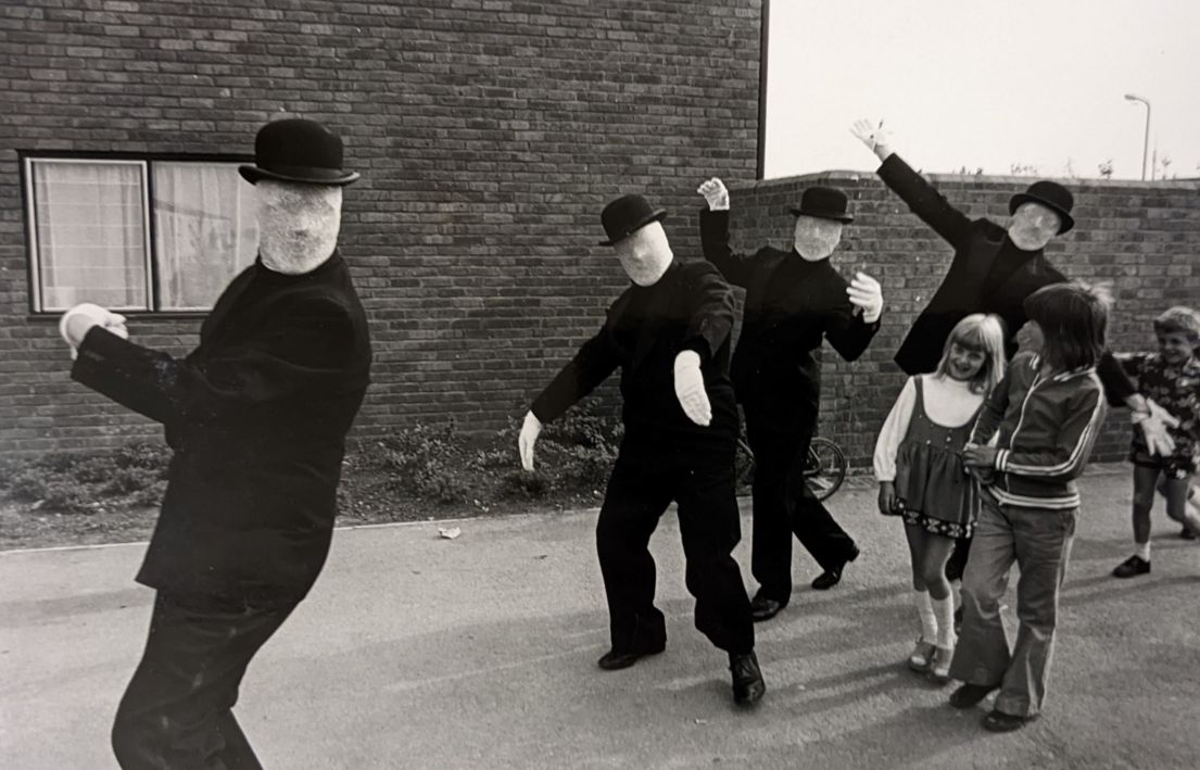 Four men in featureless masks wearing bowler hats dance-walking along a street with three children following.