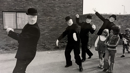 Four men in featureless masks wearing bowler hats dance-walking along a street with three children following.