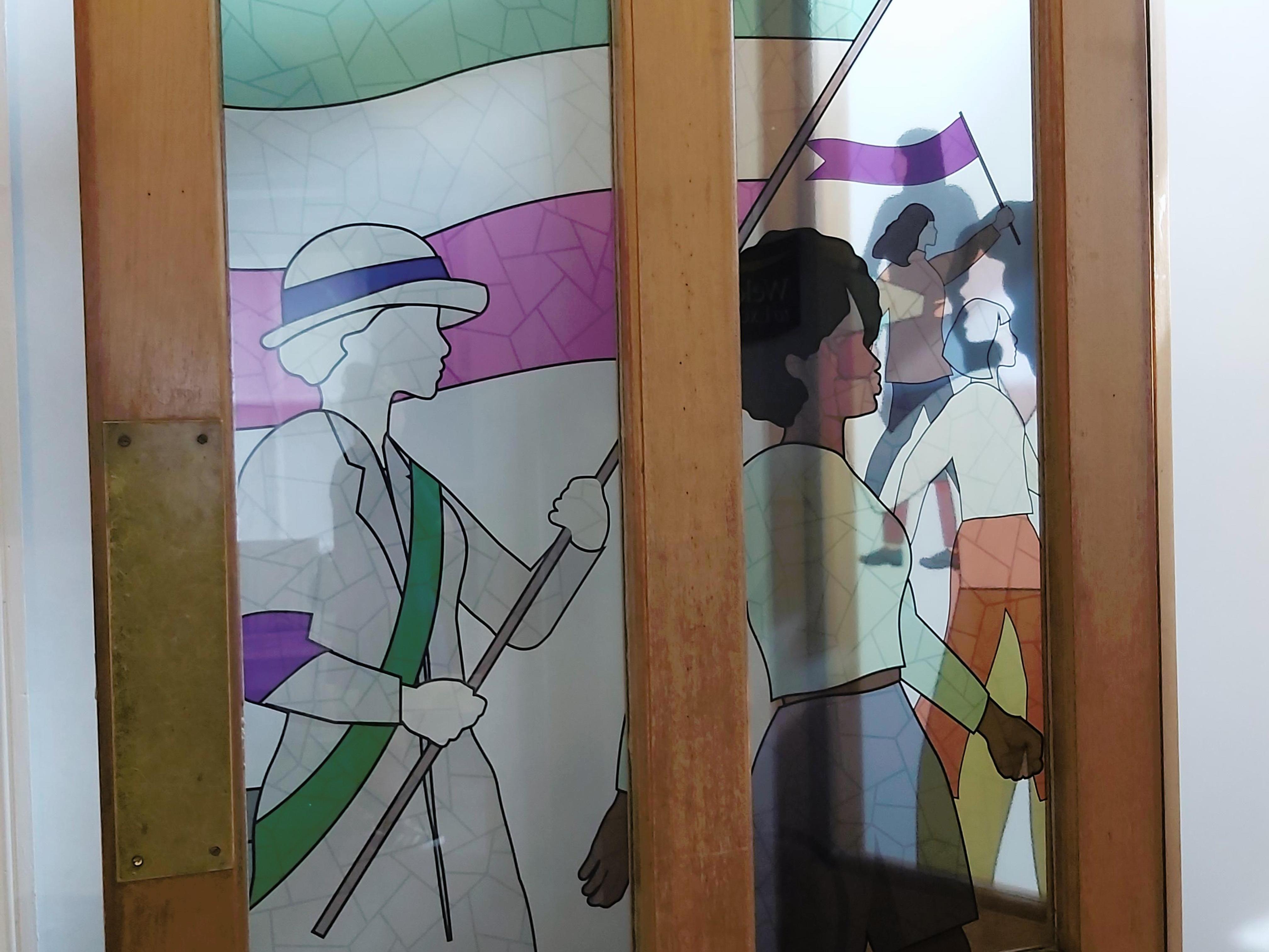 Modern stained glass door panel depicting a suffragette and then modern women in a procession.