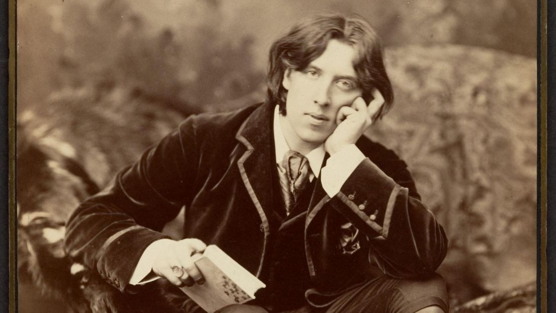 A black and white image of a gentleman with long hair, a dark waistcoat and jacket. He rests his head on one hand, in the other he holds a book.