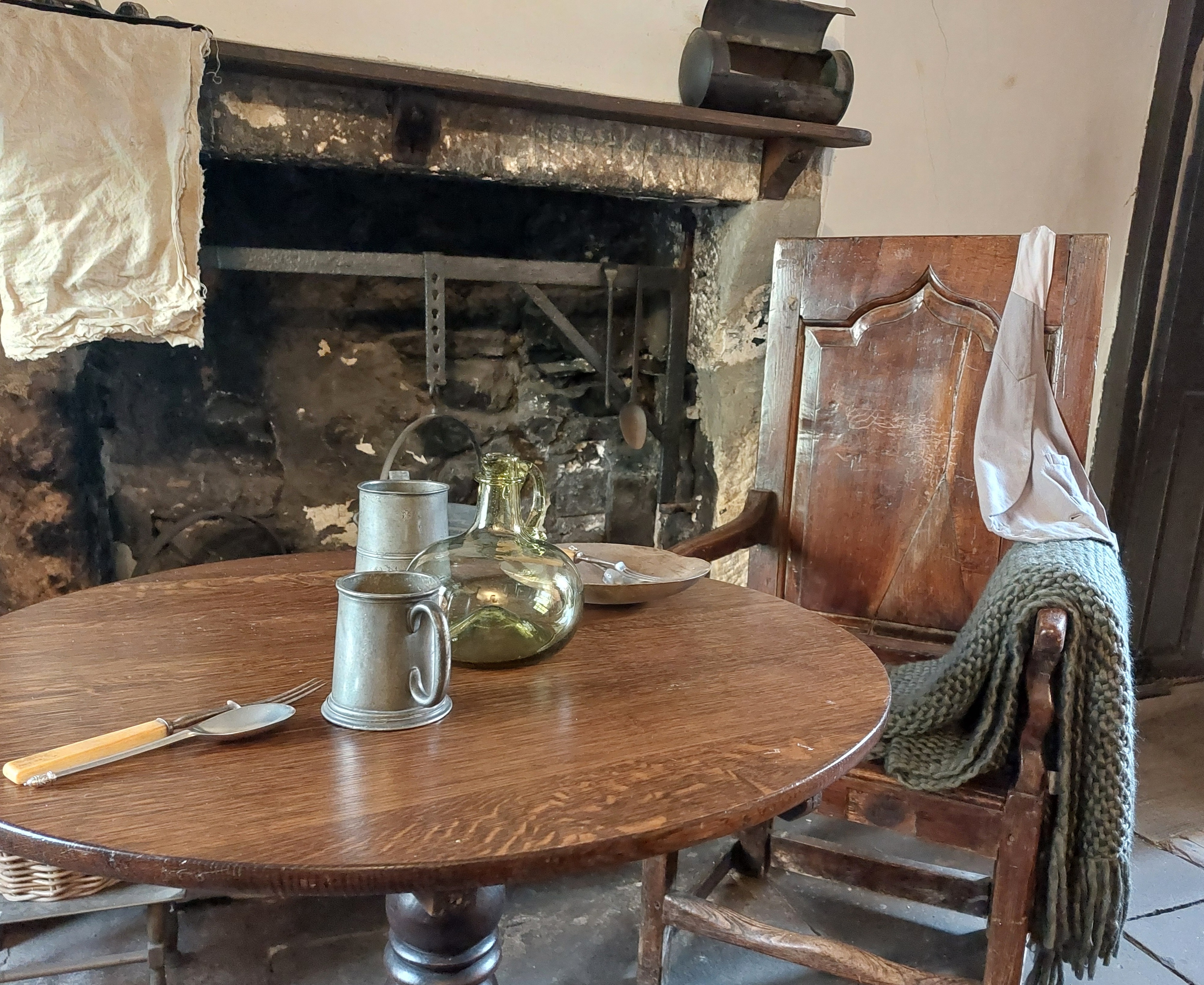 Pewter tankards and a bowl on a round wooden table and chair by a large fireplace.