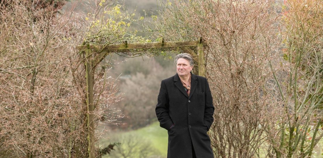 A man with grey hair, with is hands in his long black coat, walking through a wooden pagoda past some newly blossomed trees.