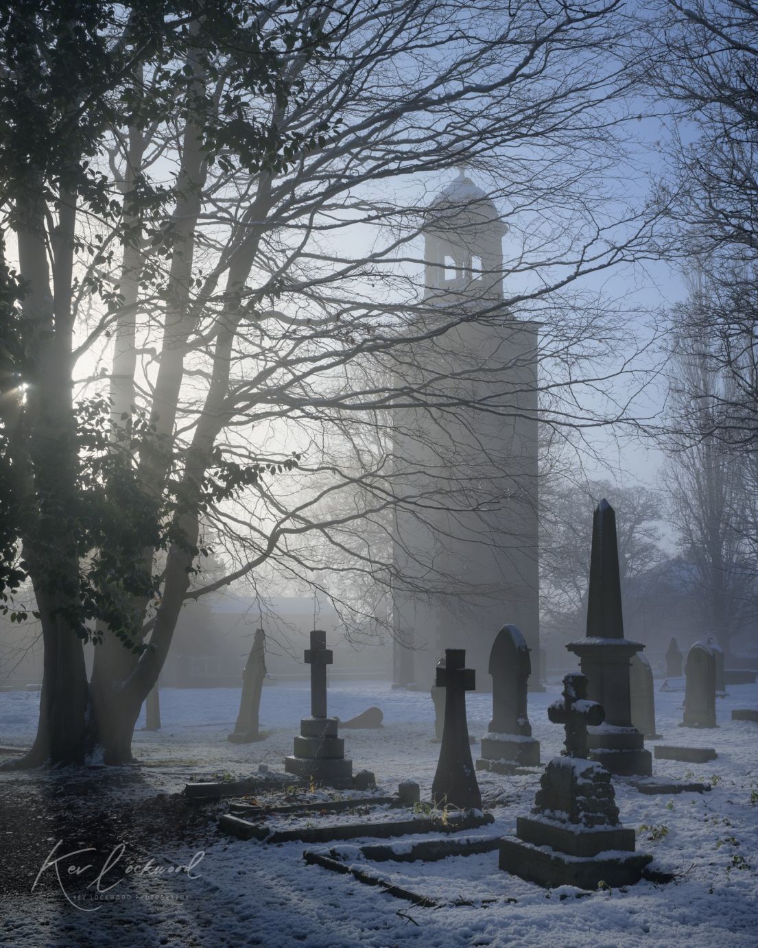 A tall silhouette of a church tower in a grave yard, surrounded by bare trees and gravestones on a frosty morning.