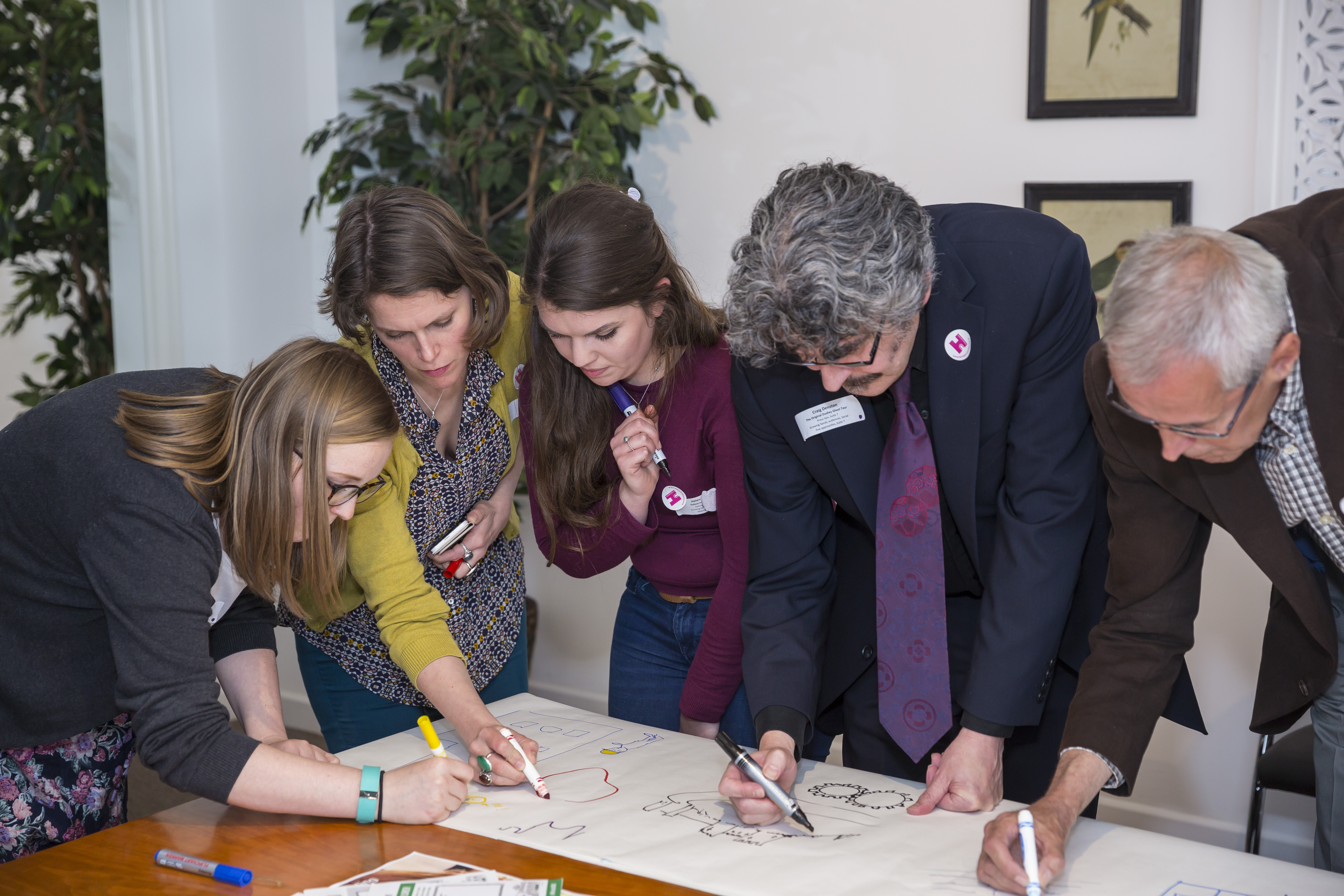 Three women and two men leaning over a table, working on a large sheet of paper.