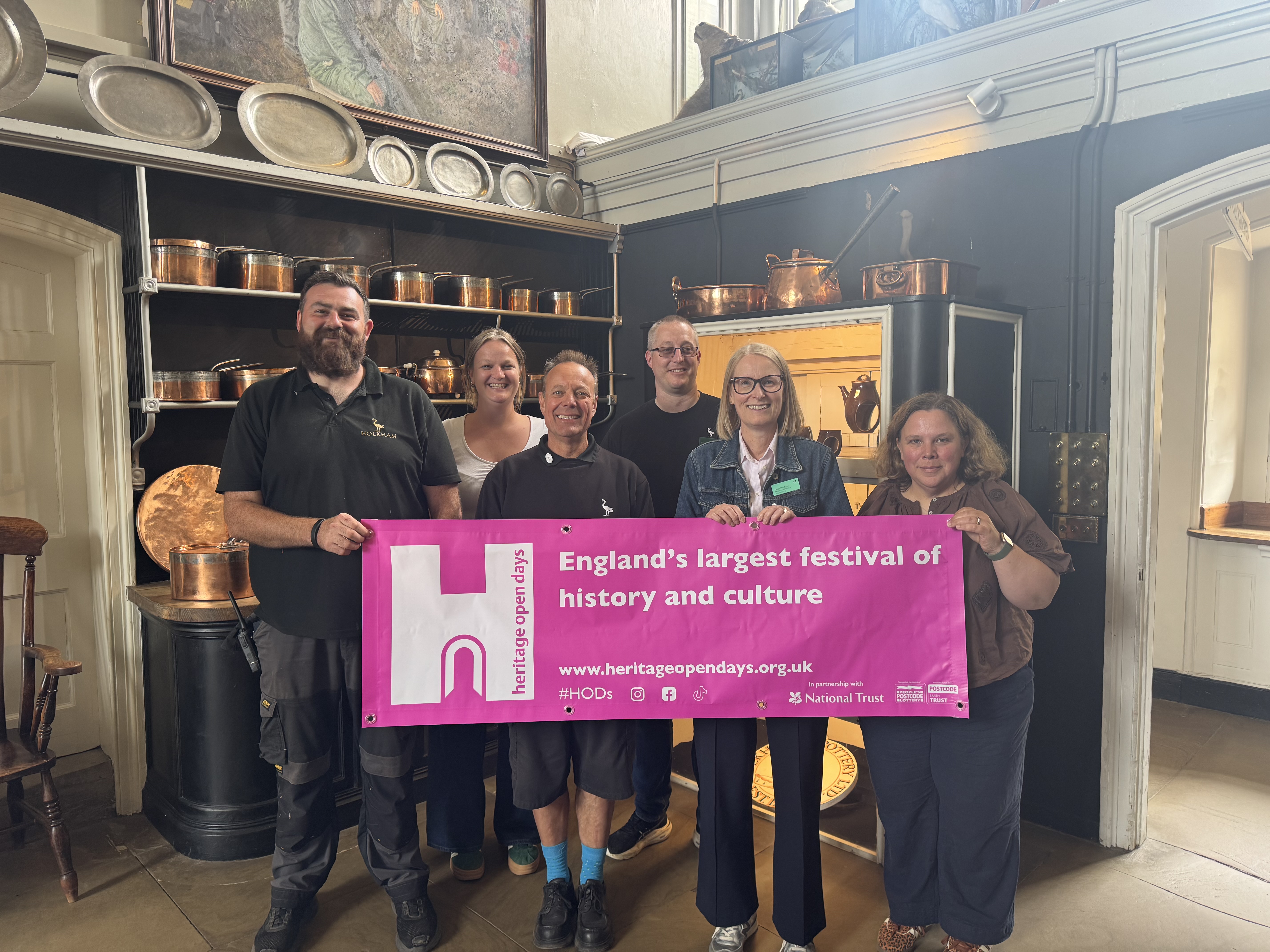 Group of people holding a banner in a kitchen with shelves of copper utensils.