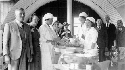 People in formal clothes gathered round a long table where women in white caps and aprons are serving food. 