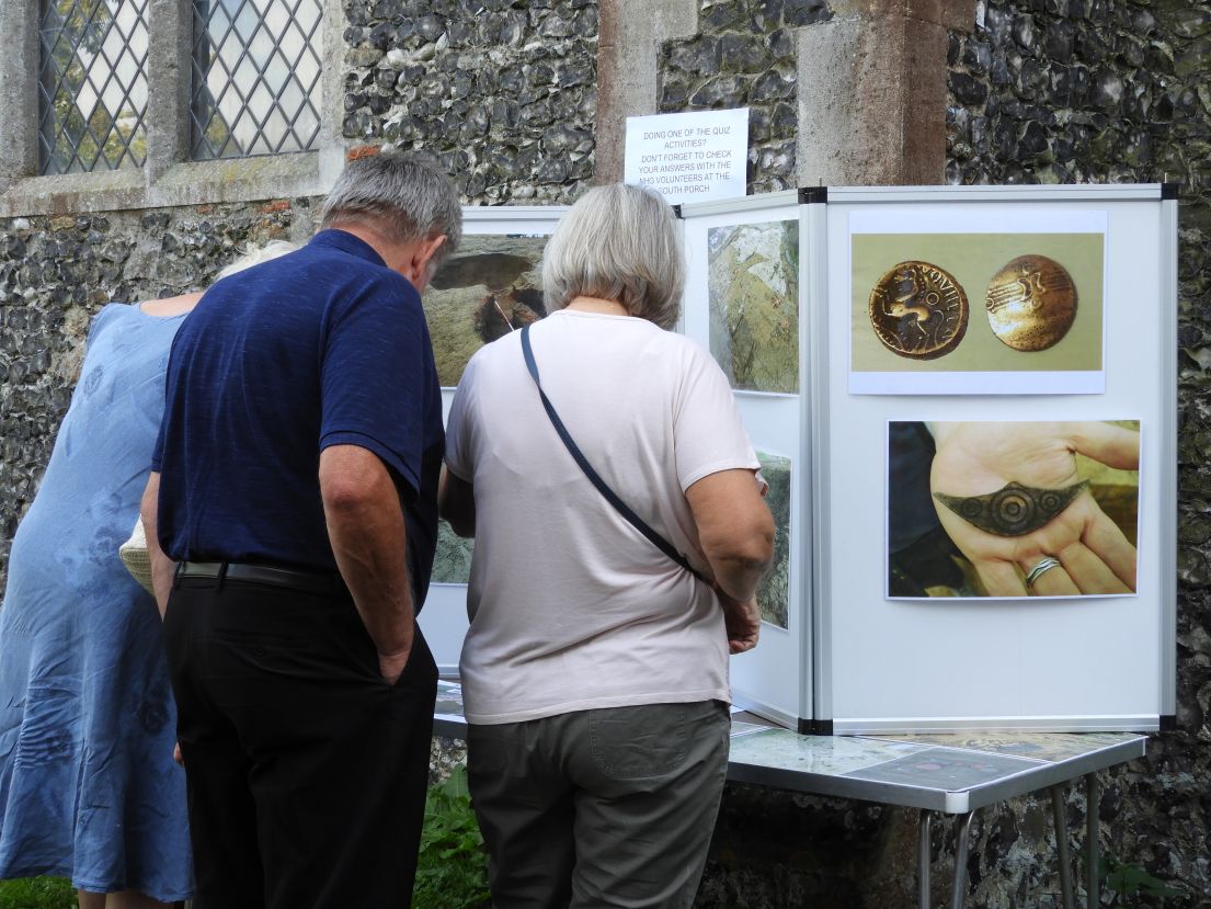 Two women and a man looking at propped up display boards with photographs. The boards on on a table outside a stone building.