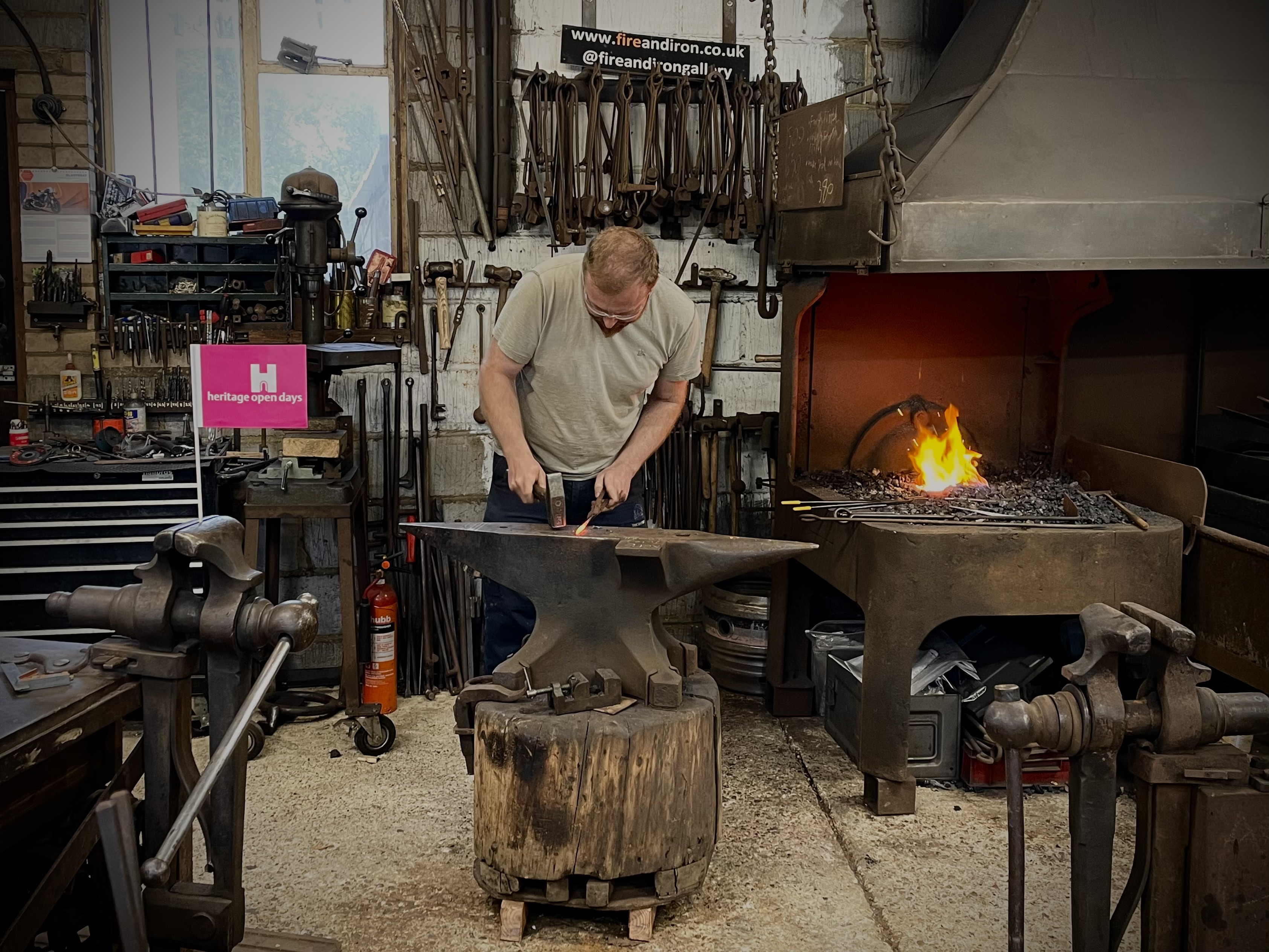 Man hammering iron on an anvil in a large room with many tools and a lit forge.