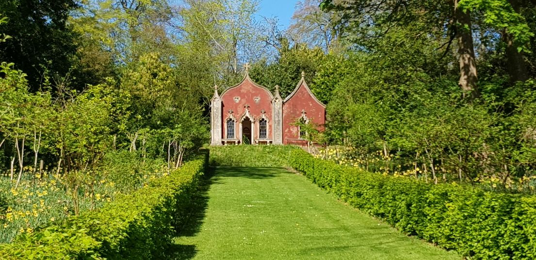 A Red brick building in the Rococo style at the end of a long garden, a path of grass defined by planting beds each side.