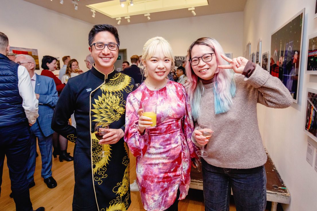 Three individuals from Queer China, dressed in bright colours and all holding drinks and toasting for a picture.