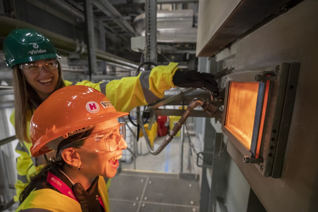 Two women wearing hard hats, one holding a HODs hand held flag and opening a viewing hatch to an incinerator. The other women looks in in amazement. 