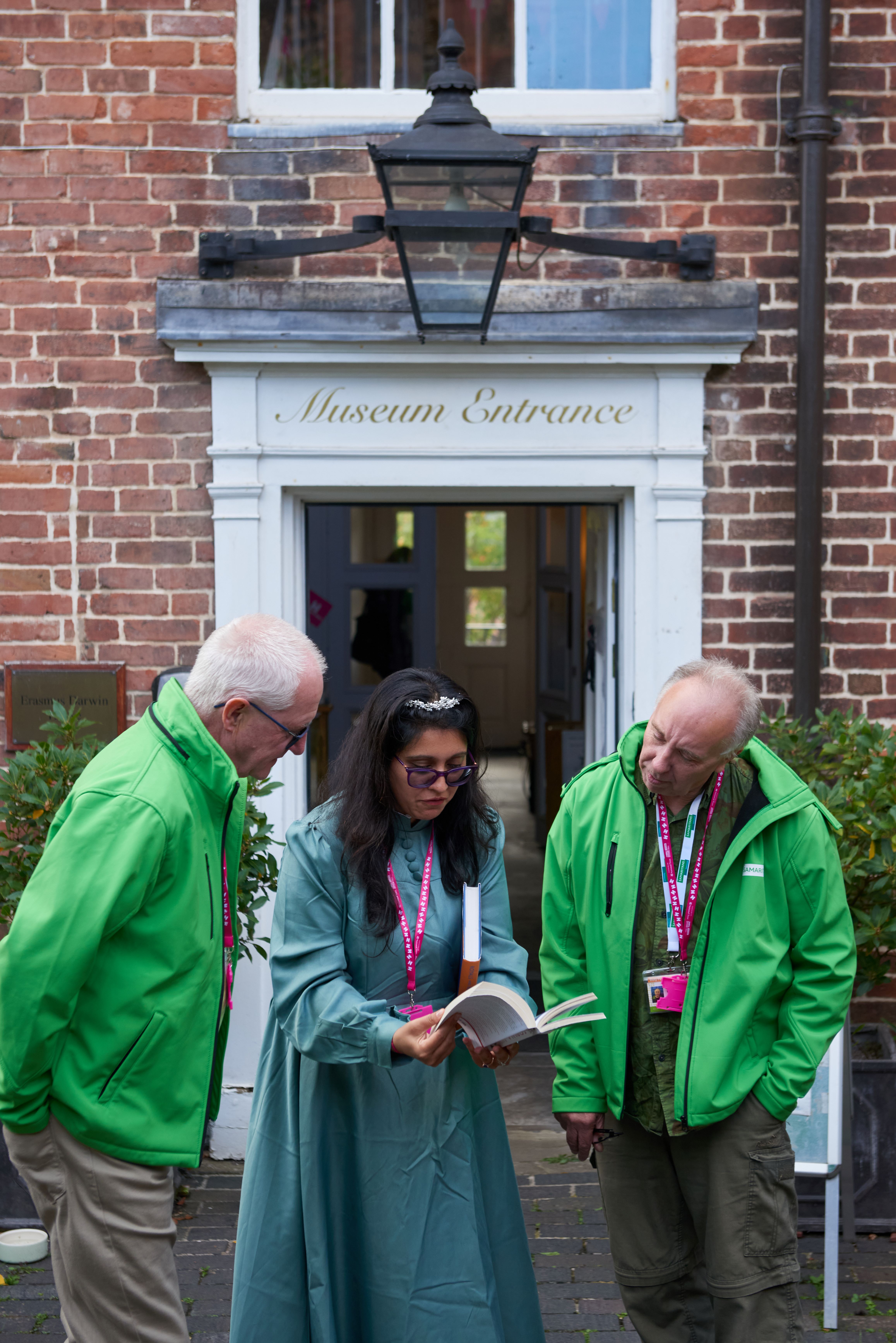 Young woman stood between two older men in green jackets showing them a book. Stood outside a museum entrance. 