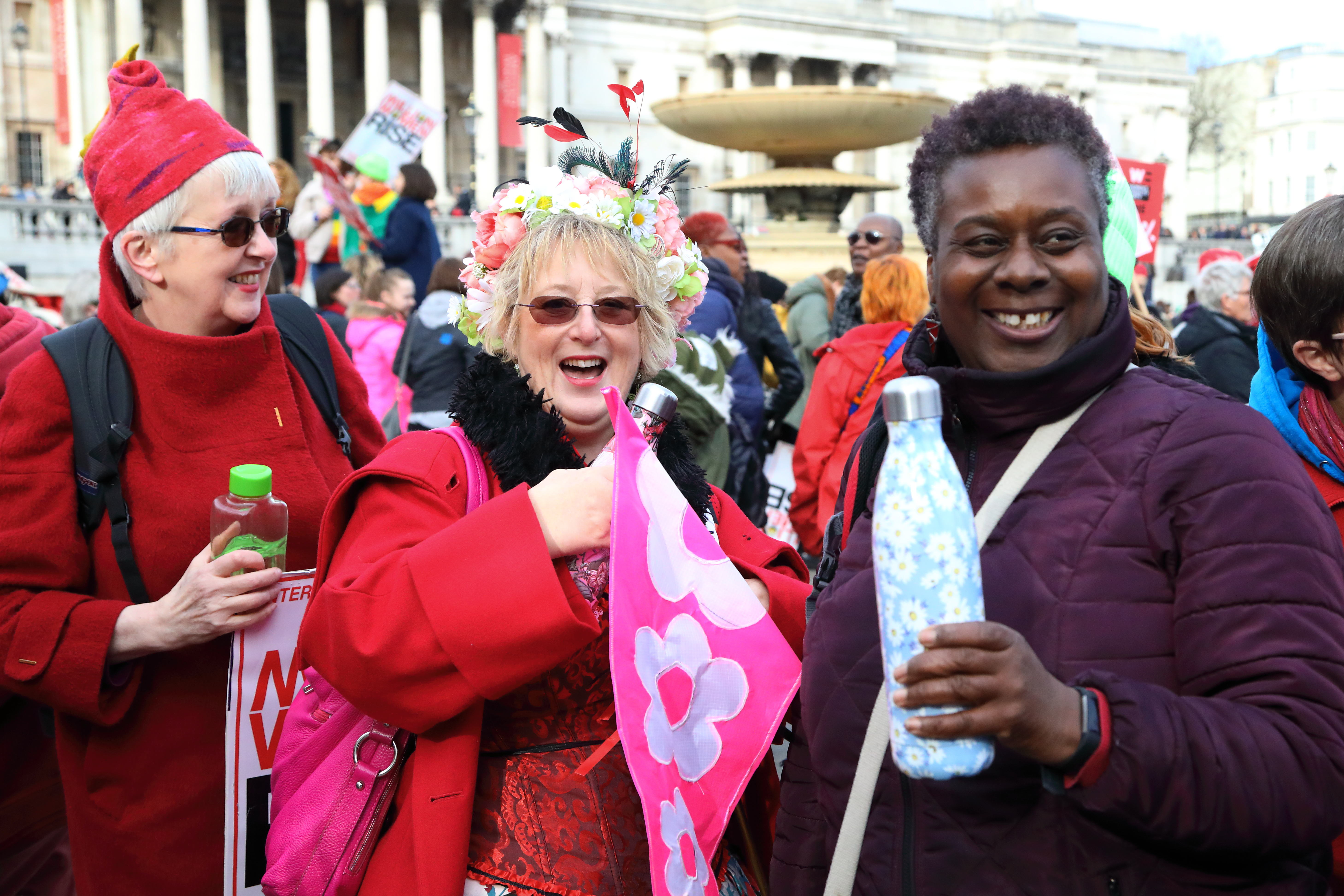 Three smiling women in warm coats, holding banners at their sides in a relaxed crowd outside a grand colonnaded building. 