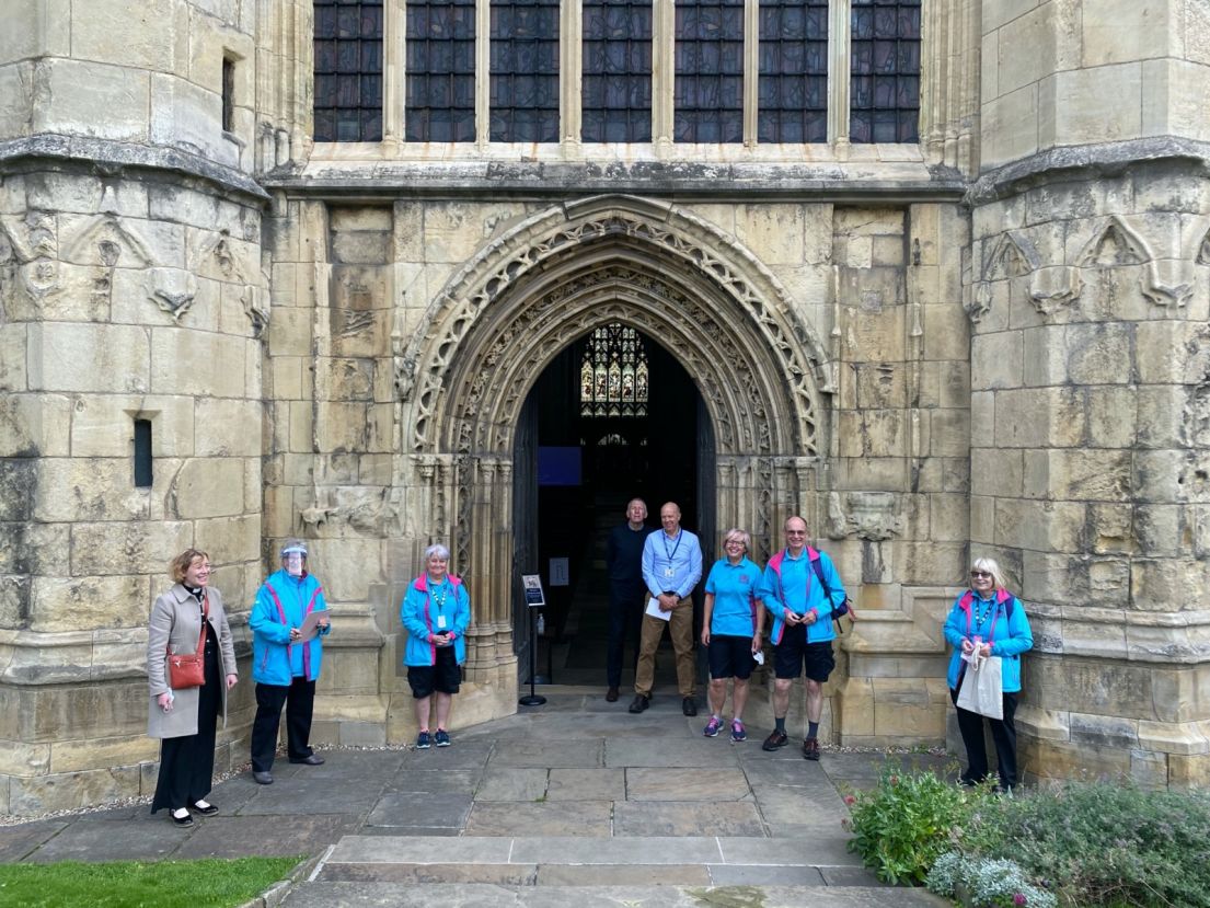 Eight people standing in front of a church decorated stone carved entrance. Five of those standing wear bright blue fleeces.