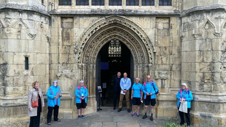 Eight people standing in front of a church decorated stone carved entrance. Five of those standing wear bright blue fleeces.