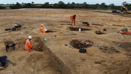 Field stripped to sandy earth with people in high vis working on small pits dug in it.
