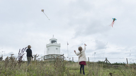 Two children playing in a grassy field with a lighthouse, both flying kites.