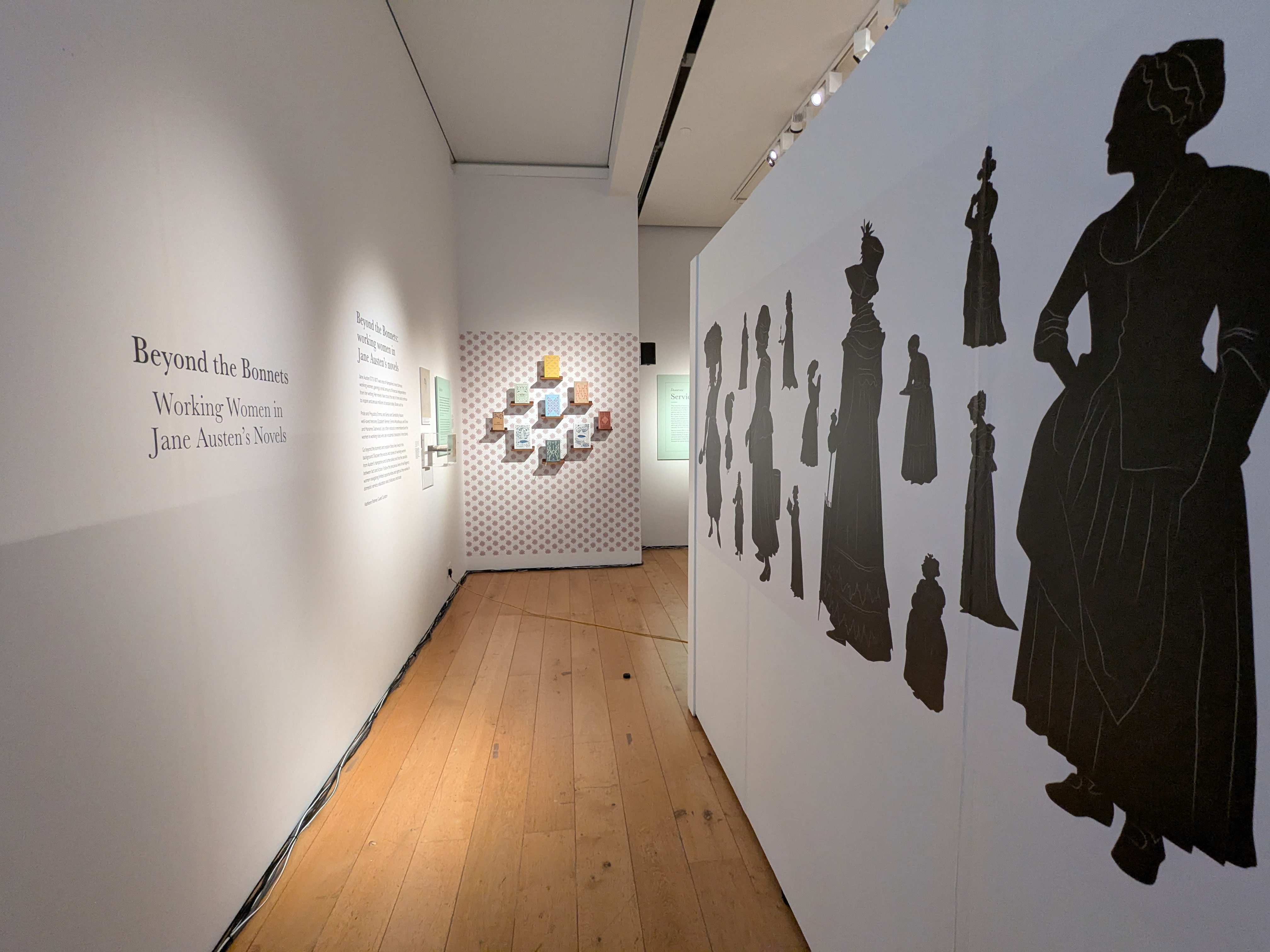 Exhibition wall with silhouettes of Georgian era women lining a short, wooden floored, corridor.