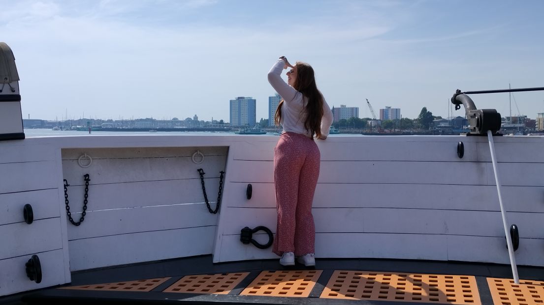 Woman stands with back to us looking out over boat rail to a view of city buildings across the water.
