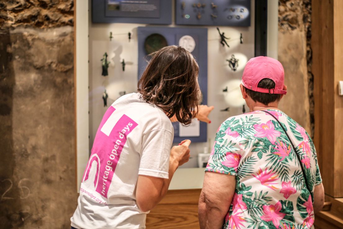 A lady with short brown hair, wearing a white top with a HODs logo on the back, talking to a visitor about a display. Both are facing away.