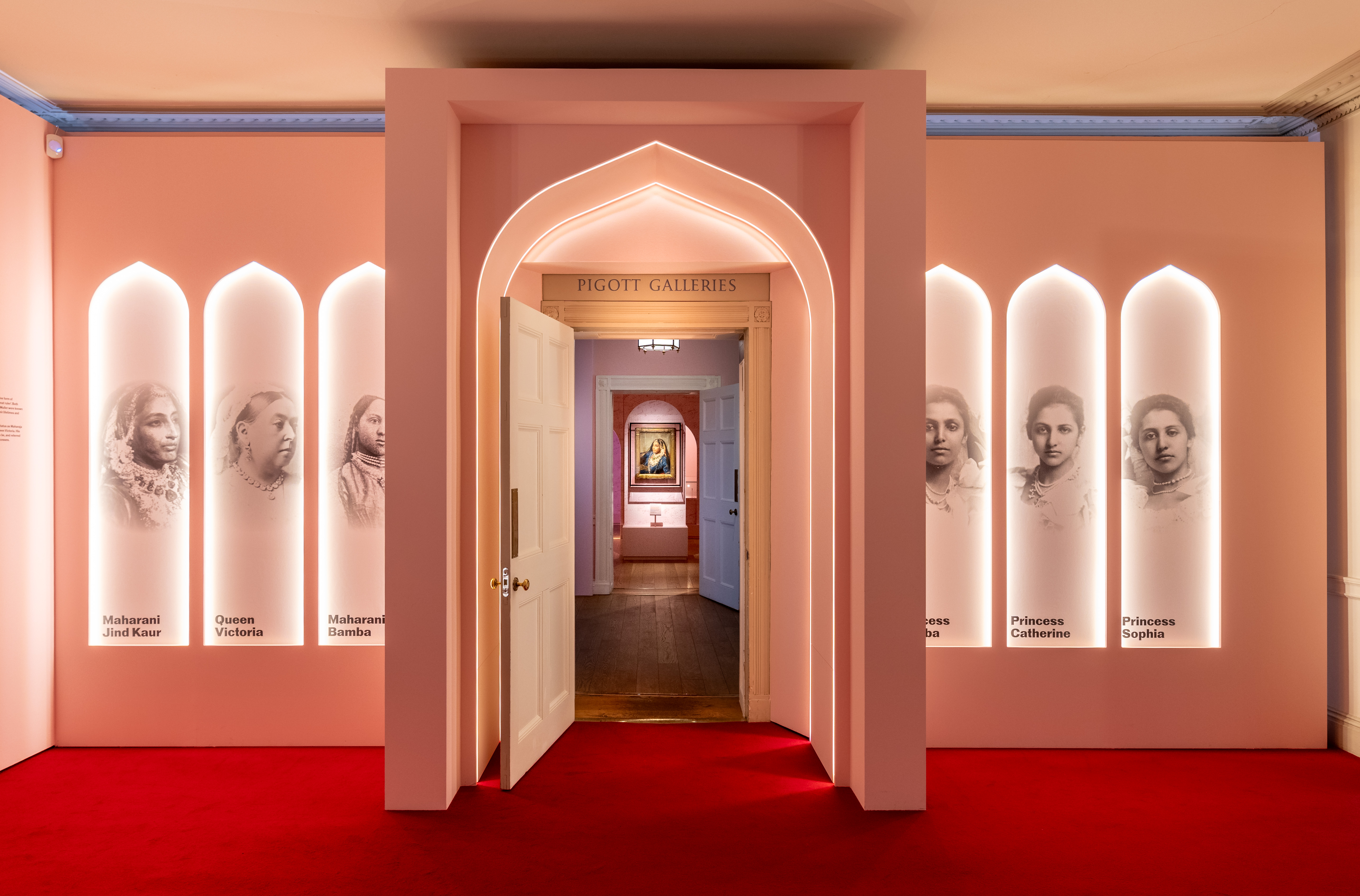 Exhibition with central arched doorway leading to a room with a spotlit painting. Arch flanked by 6 narrow arches with photos of individual women.
