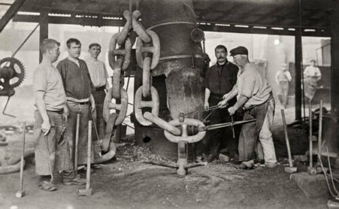 A black and white photo of 5 blacksmiths working on a gigantic chain - big enough for a cruise anchor.