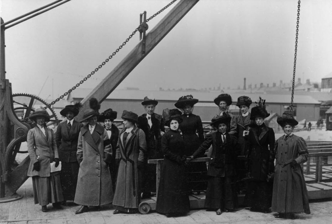 An Edwardian black and white photograph taken of women dressed in full length dark dresses, coats and hats, stood on a pier looking to the harbour.