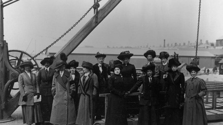 An Edwardian black and white photograph taken of women dressed in full length dark dresses, coats and hats, stood on a pier looking to the harbour.