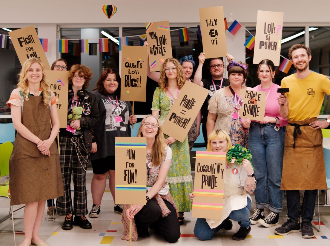 A group of people, standing in a group, holding up hand printed protest panels - with "Community Pride" and "Pride = Power" slogans.