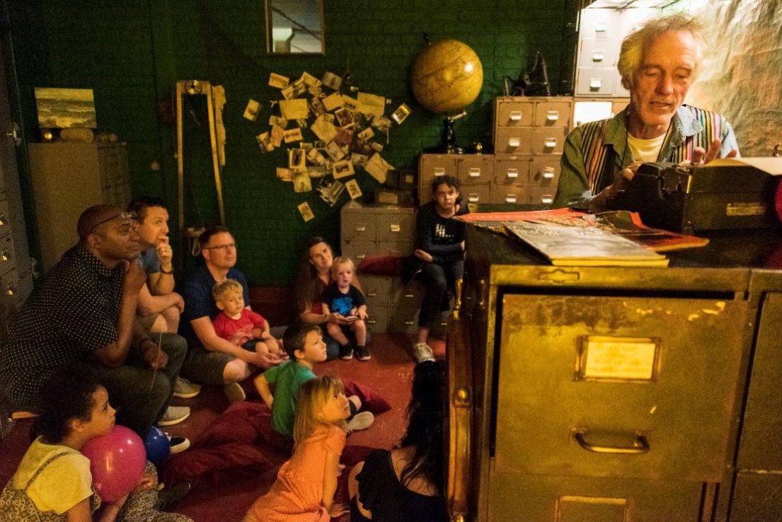 A dark room full of metal document draws. Children and their parents are sat on the floor listening intently to a man sat at a desk on a typewriter.