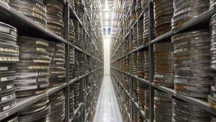 Narrow corridor in a film archive with high shelving stretching away on either side stacked with film canisters