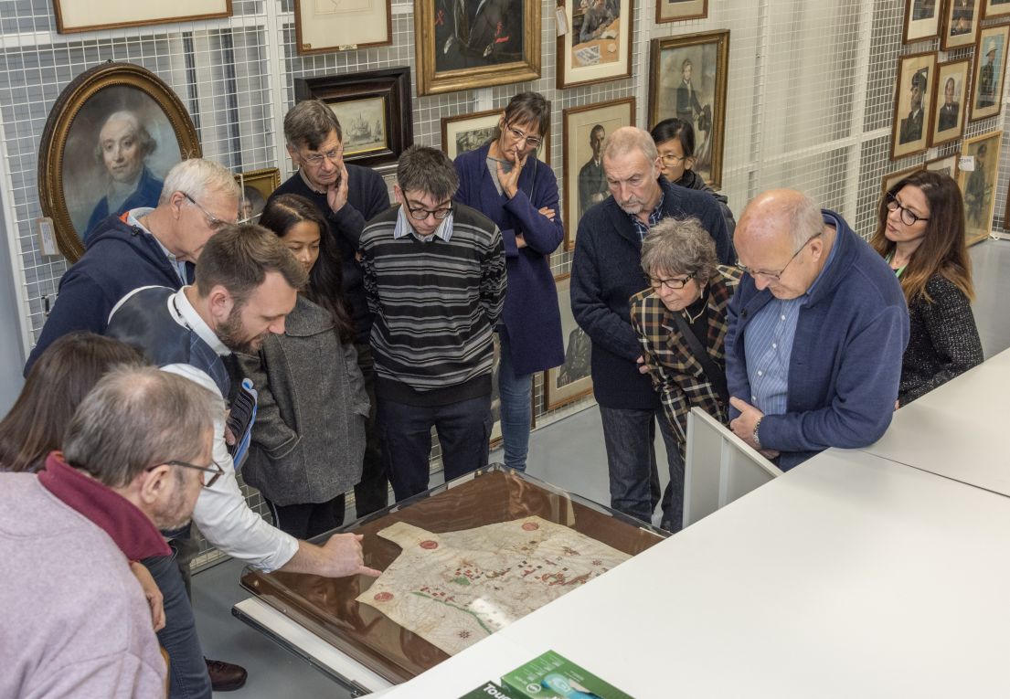 A group of people of various ages, gathered around a table with a glass draw, peering at the map enclosed inside. Behind them an array of paintings.