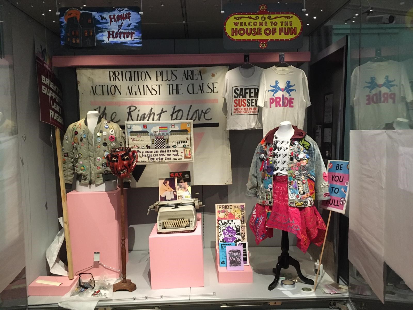 Glass museum case with pale pink plinths and an array of items including: jackets, t shirts, typewriter, leaflets.